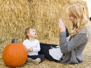 Mother photographing daughter (12-17 months) with large pumpkin