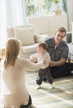 Happy parents helping little son (2-3 years) walking in living room.