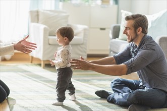 Happy parents helping little son (2-3 years) walking in living room.