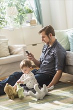 Father sitting on floor with little son (2-3 years) in living room and texting message.