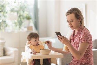 Mother feeding little boy (2-3 years) in high chair and texting message.