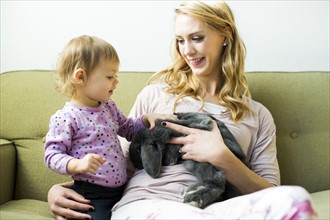 Mother and daughter (12-17 months) sitting on sofa stroking rabbit