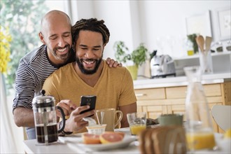 Smiley homosexual couple using smart phone at dinner table.