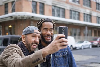 Smiley homosexual couple taking selfie with smart phone in street.