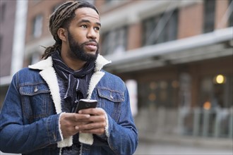 Serious man with dreadlocks holding smart phone in street.