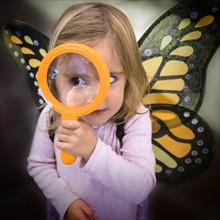 Girl (2-3) wearing butterfly wings looking through magnifying glass.