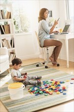 Boy (2-3) playing with toy blocks while mother working on laptop.