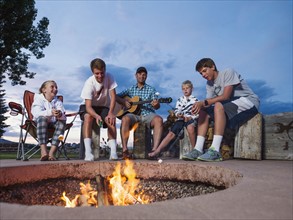 Father with children (8-9, 10-11, 16-17) sitting in front of campfire and playing guitar