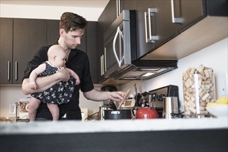 Father holding baby daughter (2-5 months) while stirring food.