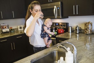 Mother drinking water while holding baby daughter (2-5 months).