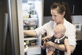 Mother looking for food in refrigerator while holding baby daughter (2-5 months).