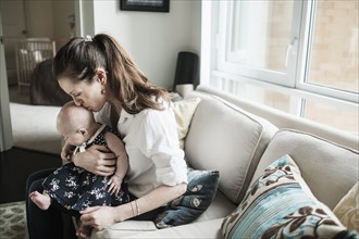 Mother kissing baby daughter's (2-5 months) head in living room.