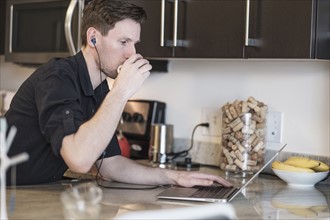 Man drinking while working on laptop computer in kitchen.