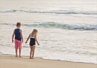 Boy (6-7) and girl (4-5) standing on beach by water