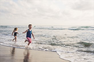 Boy (6-7) and girl (4-5) running on beach by water