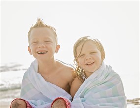 Boy (6-7) and girl (4-5) sitting on beach wrapped in towels