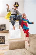 Boys (2-3, 6-7) wearing superhero costumes jumping on sofa
