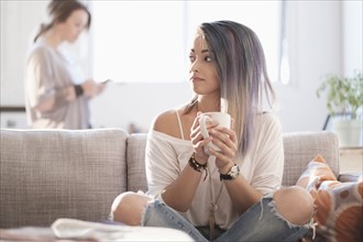 Young woman sitting on sofa and holding mug, other woman using mobile phone.
