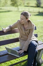 Austria, Salzburger Land, Maria Alm, Mature woman sitting on bench and using smart phone and laptop