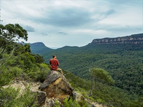 Australia, New South Wales, Katoomba, Rear view of mid adult man sitting on rock and looking at Blue Mountains