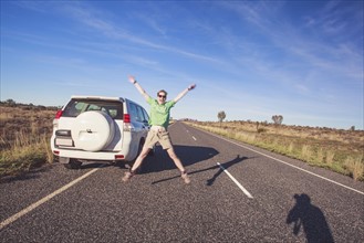 Australia, Outback, Northern Territory, Red Centre, Uluru-Kata Tjuta National Park, Woman jumping next to SUV car on road in wilderness