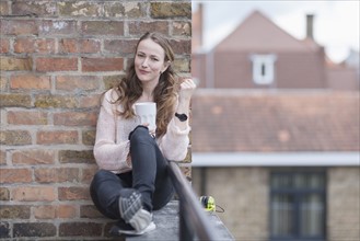 Mid-adult woman sitting on balcony railing and leaning against brick wall