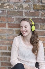 Mid-adult woman sitting and leaning against brick wall with closed eyes and headphones on