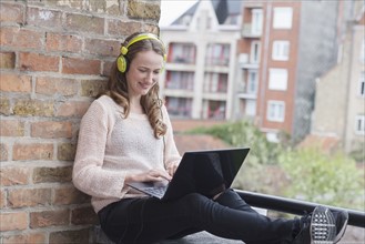 Mid-adult woman with headphones on sitting on balcony railing and using laptop