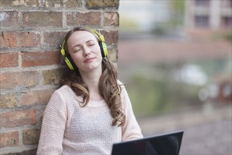 Mid-adult woman with eyes closed and headphones on leaning against brick wall