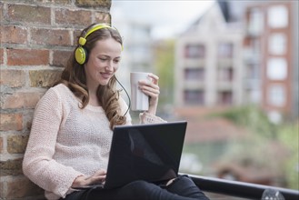 Mid-adult woman with headphones on sitting on balcony railing holding coffee cup and using laptop