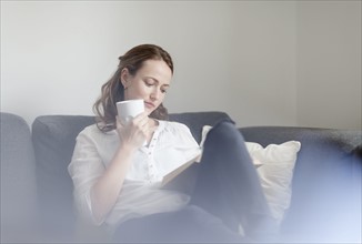Mid-adult woman sitting on sofa with book and cup of coffee