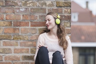 Mid-adult woman with eyes closed and headphones on sitting by brick wall