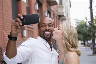 Woman kissing boyfriend in cheek, man taking selfie.