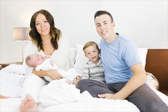 Parents sitting with daughter(0-1 months) and son (4-5) on bed in bedroom