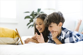 Siblings (10-11, 6-7) lying in bed and using tablet