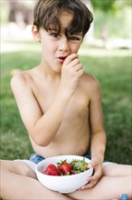 Boy (6-7) sitting on grass and eating strawberry in park