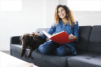 Woman sitting with dog on sofa in living room and reading book
