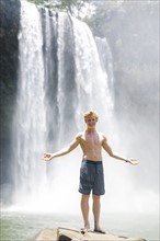 USA, Hawaii, Kauai, Young man standing on boulder in front of waterfall