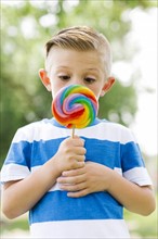 Boy (6-7) squinting and holding big, colorful lollipop