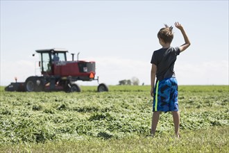 USA, Colorado, Rear view of boy (8-9) standing in field and waving to father driving combine harvester in field