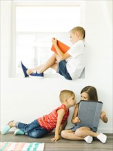 Boy (6-7) reading book and sister with brother (2-3, 6-7) using digital tablet