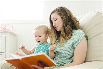 Mother with baby girl (12-17 months) sitting in living room and reading book