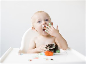Baby boy (12-17 months) sitting in high chair and eating cupcake
