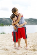 Brothers (4-5, 6-7) embracing on beach by lake