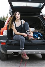 Smiling woman sitting in car trunk and tying hiking shoes