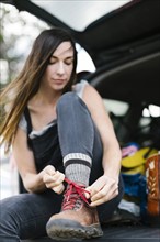 Woman sitting in car trunk and tying hiking shoes