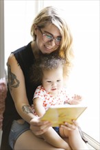 Mother sitting with daughter (12-17 months) on windowsill and reading book
