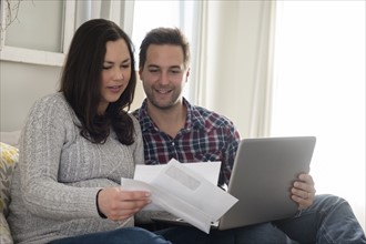 Mid adult couple sitting on sofa and looking at letter, man holding laptop