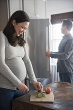 Pregnant woman cutting cucumber, man looking into refrigerator