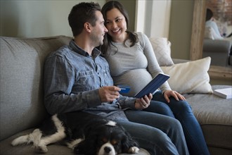 Mid adult couple shopping online in living room, sitting on sofa with puppy